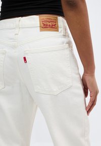 Πίσω όψη ενός ατόμου που φοράει λευκό τζιν Levi's με καφέ δερμάτινη ταμπέλα Levi's και κόκκινο λογότυπο, με το χέρι να ακουμπά στο πλευρό του.