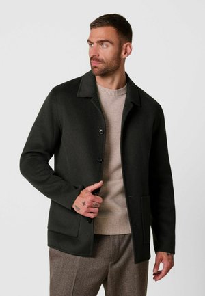 Man draagt een donker geknoopte jas over een beige trui en bruine broek, kijkt naar links met hand bij de knopen van de jas.