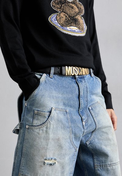 Suéter negro con un gráfico de un oso de peluche, combinado con pantalones de mezclilla azul claro desgastados y un cinturón negro con lettering dorado "Moschino".