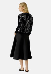 Zwarte pailletten sweater met lange mouwen, gecombineerd met een vloeiende zwarte rok. Model draagt zwarte haksandalen, die een gladde textuur laten zien.