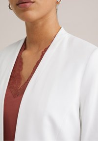 Valkoinen bleiseri, jossa sileä pinta, yhdistetty viininpunaiseen toppiin, jossa pitsireunus kaula-aukolla. Yksityiskohdat korostavat kontrastia ja kerrostamista.