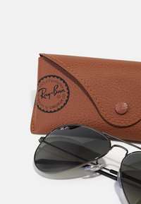 Ochelari de soare Ray-Ban aviator cu lentile întunecate, cadru din metal și suporturi pentru nas reglabile, lângă un toc din piele maro texturată cu logo.