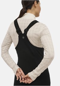 Sort bib med justerbare stropper, tekstureret materiale på ryggen, over en lys mønstret langærmet bluse. Hænderne knyttet bag på.