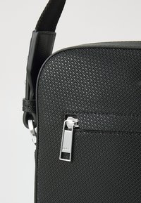Borsa in pelle nera con texture, superficie liscia, tasca frontale con zip e tracolla removibile con finiture in metallo.