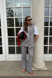 Gilet e pantaloni grigi chiari su misura, abbinati a una t-shirt bianca. Gli accessori includono una borsa nera con accenti rossi e infradito rossi.