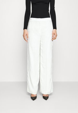 Persoon die witte wijde broek met hoge taille draagt, een zwart nauwsluitend topje met lange mouwen en zwarte puntige hakken, staand tegen een effen achtergrond.