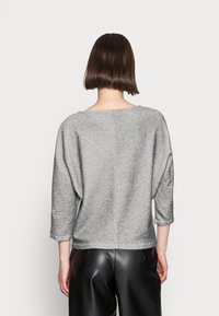 Harmaa, tekstuuriinen ylioversize-neule, jossa on kolmen neljänneksen hihat, leveä kaula-aukko ja pystysauma selkäpuolella. Yhdistetty mustiin nahkahousuihin.