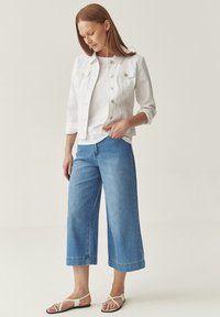 Veste en denim blanche avec boutons, associée à un haut blanc et à un jean bleu clair à jambes larges. La mannequin porte des sandales à lanières avec des fines lanières.