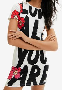 Kratší šatové šaty z bílé látky s výrazným černým textem a červenými květinovými akcenty. Úzký střih s kulatým výstřihem a krátkými rukávy.