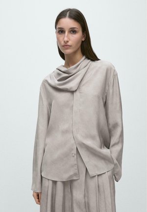 Kvinde med langt brunt hår iført løs, lysegrå langærmet top og matchende plisseret nederdel mod ensfarvet baggrund.