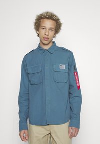 Modrá košeľa na gombíky vyrobená z bavlnenej zmesi, s dvoma náprsnými vreckami, ramennými epauletmi a červeným zipsovým vreckom s textovým detailom.