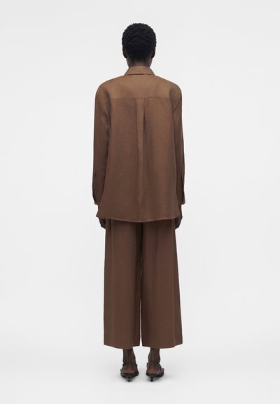 Άτομο με κοντά σγουρά μαλλιά φοράει χαλαρό καφέ μακρυμάνικο πουκάμισο και φαρδιά παντελόνια, στέκεται γυρισμένο με την πλάτη σε απλό φόντο.