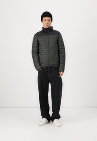 Polstret grøn jakke med sort forkant, lynlås lukning og ståkrave, parret med loose black bukser og hvide sneakers.