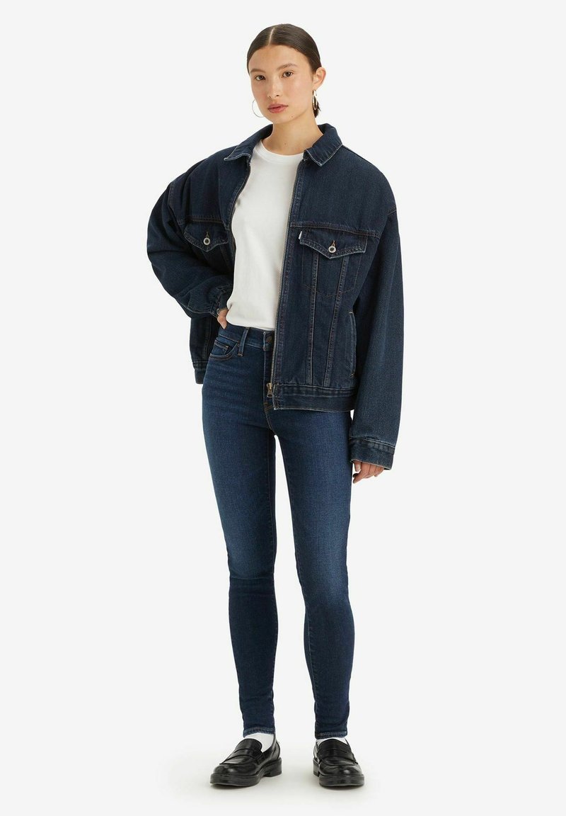 Chaqueta de mezclilla azul oscura de corte oversize, con dos bolsillos frontales y una camiseta blanca debajo. Combinada con jeans oscuros de corte slim y zapatos negros.
