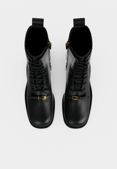Paire de bottes montantes en cuir noir avec bouts carrés arrondis, lacets à l’avant, boucles dorées et tirettes de fermeture éclair latérales, vues de dessus.