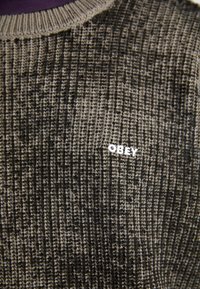 Pletene pulover v temno sivi in črni barvi, ki ima teksturiran vzorec in majhen bel logotip "OBEY" na prsih. Okrogla izrezana vrata.