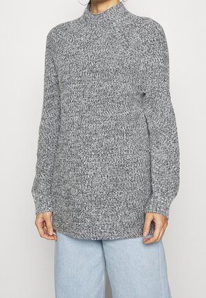 Personne portant un pull en maille gris à col montant et un jean bleu clair, debout devant un fond blanc uni.