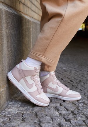 Persoon draagt beige broek en wit-roze Nike hoge sneakers, staand op oneffen kasseien naast een stenen muur.