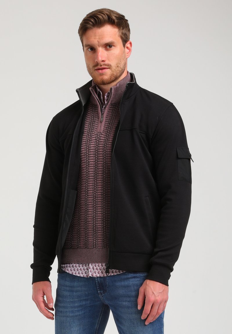 Giacca nera con zip e texture a costine, maniche lunghe e tasche laterali, indossata sopra una camicia a fantasia con colletto alto.