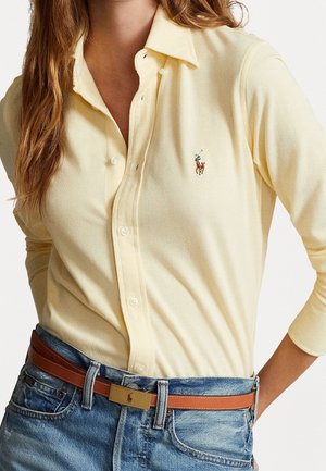 Vrouw die een lichtgele blouse met knopen draagt met een klein paardenpolo-logo en blauwe spijkerbroek met een bruine leren riem met een gouden gesp.