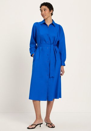 Frau in einem langärmeligen, blauem Midikleid mit Knopfleiste und gebundenem Taillengürtel, die mit den Händen in den Taschen steht und schwarze Sandalen mit Absatz trägt.