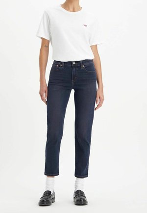 Personne portant un t-shirt blanc, un jean taille haute bleu foncé, des chaussettes blanches et des mocassins noirs, debout contre un fond uni.