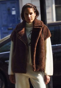 Puszysta brązowa kamizelka bez rękawów z zamkiem z przodu i kołnierzykiem, noszona na kremowym swetrze. Miękka faktura i luźny krój.