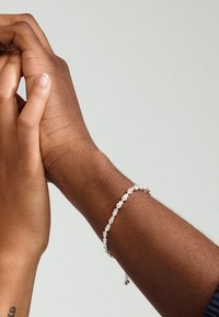 Pulsera de plata con piedras transparentes en forma circular, descansando sobre un brazo de piel oscura, mientras una mano de piel clara la toca suavemente. Fondo neutro.