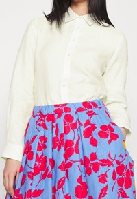 Camicia di lino verde chiaro a maniche lunghe con colletto abbottonato, abbinata a una gonna blu con motivi floreali rossi e una vita elasticizzata.