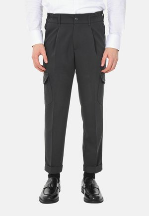 Pantaloni cargo neri su misura con tasche laterali, indossati con mocassini neri con nappine e una camicia bianca su una persona in piedi.
