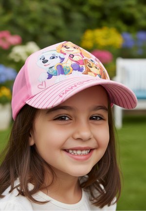 Smiling young girl wearing a pink cap with colorful animated puppies, outdoors with grass and blurred flowers in the background.
