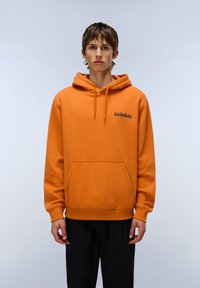 Felpa arancione realizzata in materiale morbido, con una tasca frontale, cappuccio regolabile e un piccolo logo nero sul petto. Abbinata a pantaloni neri.
