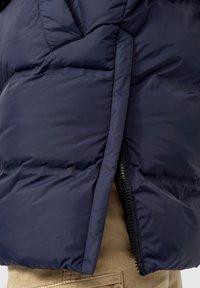 Gros plan sur une veste matelassée bleu marine partiellement zippée portée sur un pantalon beige en velours côtelé.
