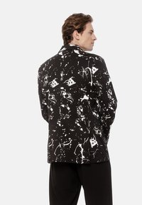 Mężczyzna w czarnym żakiecie z białym wzorem abstrakcyjnych plam farby, stojący z rękami w kieszeniach, zwrócony bokiem w stronę jednolitego tła.