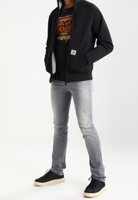 Sudadera negra con cremallera y bolsillos, camiseta gráfica gris debajo, jeans slim-fit gris claro y zapatillas negras con suelas beige.