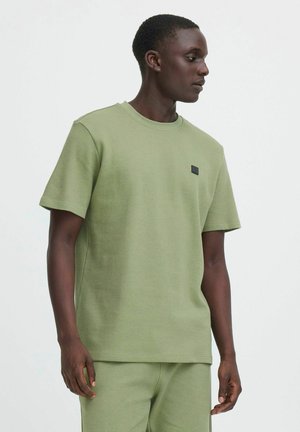 Mann trägt ein hellgrünes, strukturiertes Kurzarmhemd und dazu passende Hose, schaut seitlich vor einfarbigem Hintergrund.