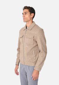 Giacca in misto lana beige con zip frontale, due tasche sul petto, colletto e maniche lunghe, caratterizzata da una texture liscia e una vestibilità su misura.