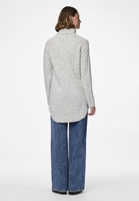 Sieviete ar īsiem brūniem matiem, tērpusies gaiši pelēkā augstu kakla džemperī un platām zilām džinsu biksēm, stāv ar muguru pret baltu fona sienu.