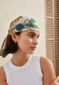 Pañuelo de seda con patrones florales en azul, amarillo y verde. Anudado en la parte trasera, complementado con una blusa blanca sin mangas.