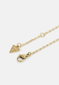 Gold-colored chain featuring a triangular pendant with "GUESS" engraving and a lobster clasp. Sleek links and a shiny texture.