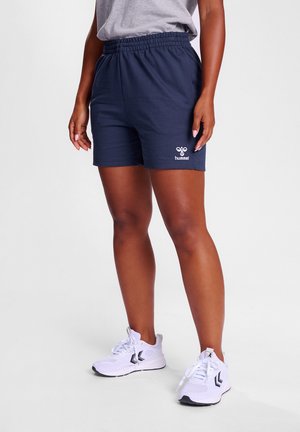 Person trägt marineblaue Sportshorts mit Hummel-Logo, graues Shirt und weiße Sneaker mit schwarzen Chevron-Akzenten.
