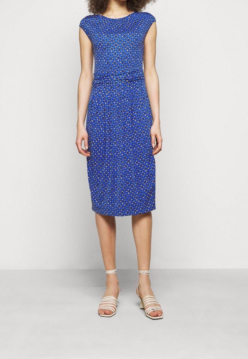 Femme portant une robe bleue à motifs géométriques longueur genou avec des manches courtes et des sandales blanches à talons blocs, debout sur un fond blanc.
