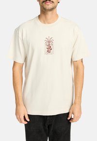 Camiseta de algodón color crema de manga corta con un gráfico central en rojo oscuro, que representa un diseño de serpiente y palma con acentos de texto decorativos.