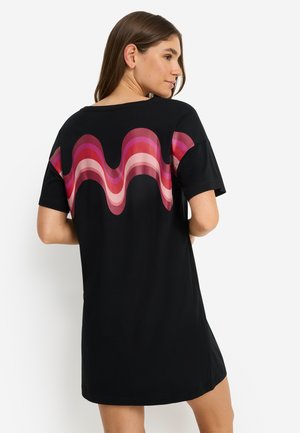 Žena u crnoj haljini kratkih rukava s uzorkom valovitih pruga u ružičastoj i crvenoj boji preko leđa, stoji na bijeloj pozadini.