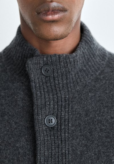 Pull en tricot gris foncé avec un col côtelé, comportant deux boutons noirs sur le devant. Texture lisse, design ajusté.