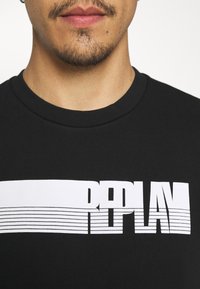 Camiseta de algodón negra con un gran logotipo gráfico blanco y detalles de rayas en el pecho, cuello redondo y mangas cortas.