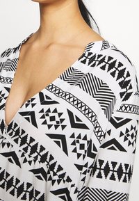 Blouse à motifs noir et blanc avec un col en V, présentant des designs géométriques et un tissu texturé, mettant en avant des formes audacieuses et contrastées.