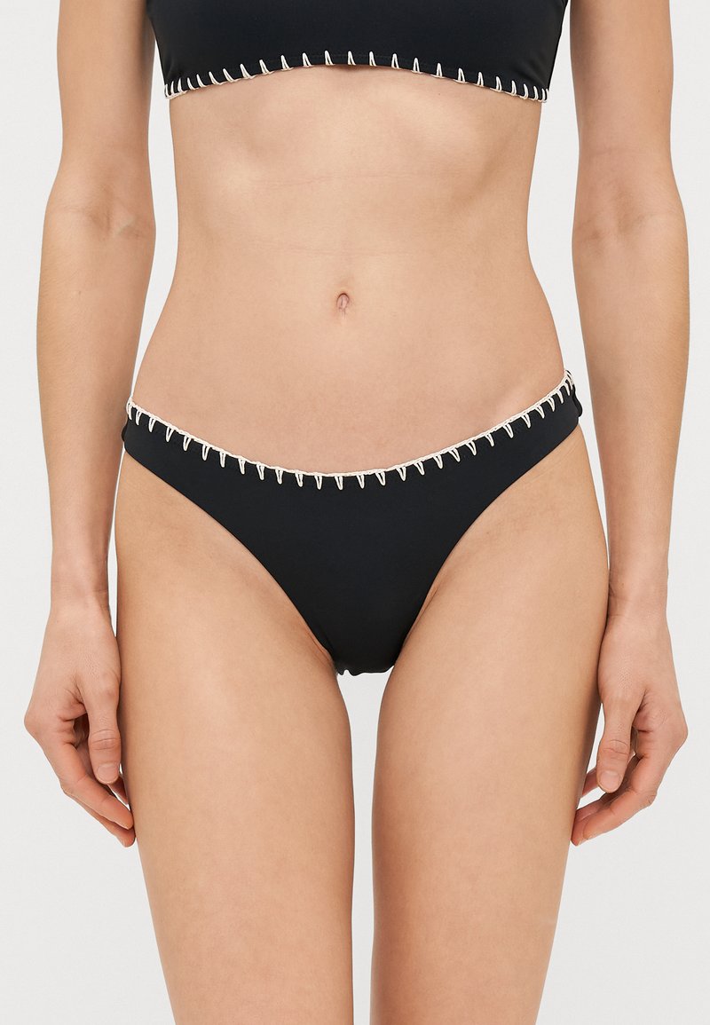 Juodas bikini dugnas su kontrastinga kreminės spalvos siuvimo linija aplink juosmenį. Ypatinasi žemu kirpimu ir lygiu tekstūru.