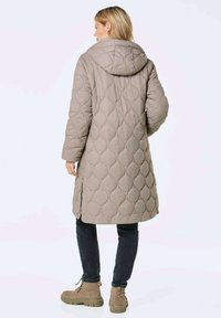 Gepolsterter langer Mantel in Taupe mit Kapuze, mit einem Rauten-Muster und seitlichen Schlitzen. Kombiniert mit dunklen Jeans und beigen Ankle Boots.