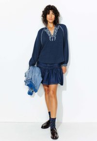 Blouse en coton bleu marine avec des détails brodés, associée à une jupe en denim. Les accessoires comprennent une veste en denim clair et des chaussettes à motifs.